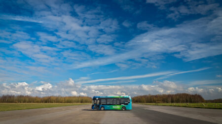 An autonomous vehicle stands parked across a test track under blue skies with grass on either side and a tree line in the distance