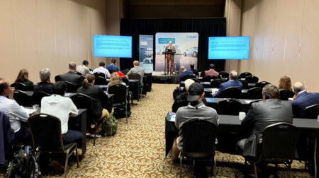 ASAM workshop participants sit in rows in a conference room with two large screens at the front and a person on a stage giving a presentation. The walls and carpet are both grey.