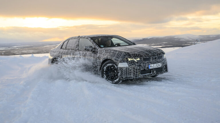 The BMW Neue Klasse i3 electric vehicle drives across a snow-covered road in Arctic cold weather endurance testing. The sun is low in the sky sand there is dominant cloud cover.