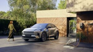 A silver Toyota car charging outside a house with wooden paneling, surrounded by greenery.
