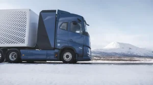 A Volvo Trucks hydrogen ICE heavy truck with a dark blue cab and grey trailer undergoes testing in a rural location with snow on the ground and a mountain visible in the background.