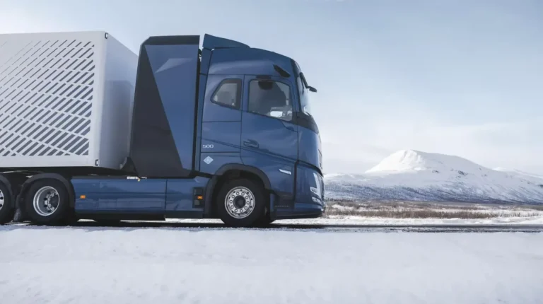 A Volvo Trucks hydrogen ICE heavy truck with a dark blue cab and grey trailer undergoes testing in a rural location with snow on the ground and a mountain visible in the background.