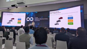 A curved sc reen displays the VI-Grade Smart Prototypes Summit logo to a room full of conference attendees.