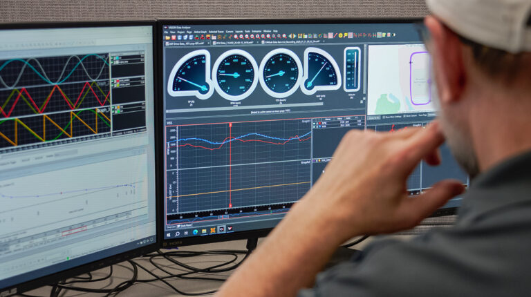 A man wearing a grey baseball cap sits in front of a pair of computer monitors displaying complex design and testing information.