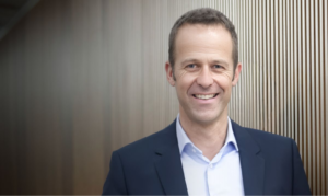 Promo photo of Joost Kessels, chief development officer of SEAT and Cupra. He is wewaring a white shirt and a dark blue suit jacket and stands in front of a dark wood wall.