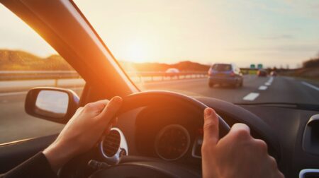 A driver's hands on the wheel of a vehicle with the sun setting in the distance through the windshield.