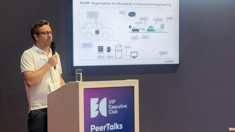 Vehicle Tech Week: ‘Standardization in the age of AI’ roundtable highlights risks and opportunities A man wearing a white shirt stands at a lectern holding a microphone giving a speech to a room of conference attendees.