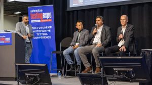 Conference speakers sit on stage at Automotive Testing Expo North America, with a conference host standing side stage at a lectern.