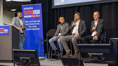 Conference speakers sit on stage at Automotive Testing Expo North America, with a conference host standing side stage at a lectern.