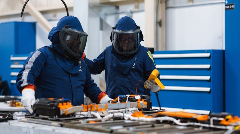 Two laboratory workers wearing blue protection suits and visors inspect a piece of equipment.